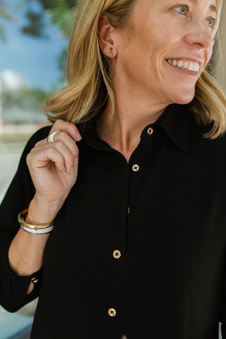 A close-up lifestyle shot of a woman wearing a cotton stretch black button-down dress with gold buttons. She is smiling and looking to the side, with a blurred outdoor background.