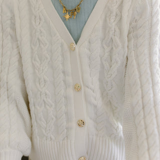 A close-up of a person wearing a 100% cotton white cable-knit cardigan over a light blue top, with gold necklaces visible against a softly blurred indoor background.