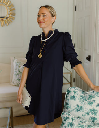 A woman wearing a navy blue puff-sleeve dress and layered gold and pearl necklaces, holding a white clutch in a bright, modern living room.