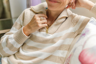 A close-up lifestyle shot of a person wearing a beige and white striped knit quarter-zip sweater, adjusting a gold necklace.